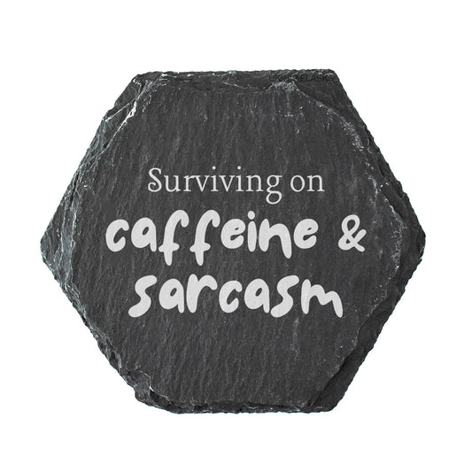 Hexagonal black slate coaster with 'Surviving on caffeine & sarcasm' text on a white background
