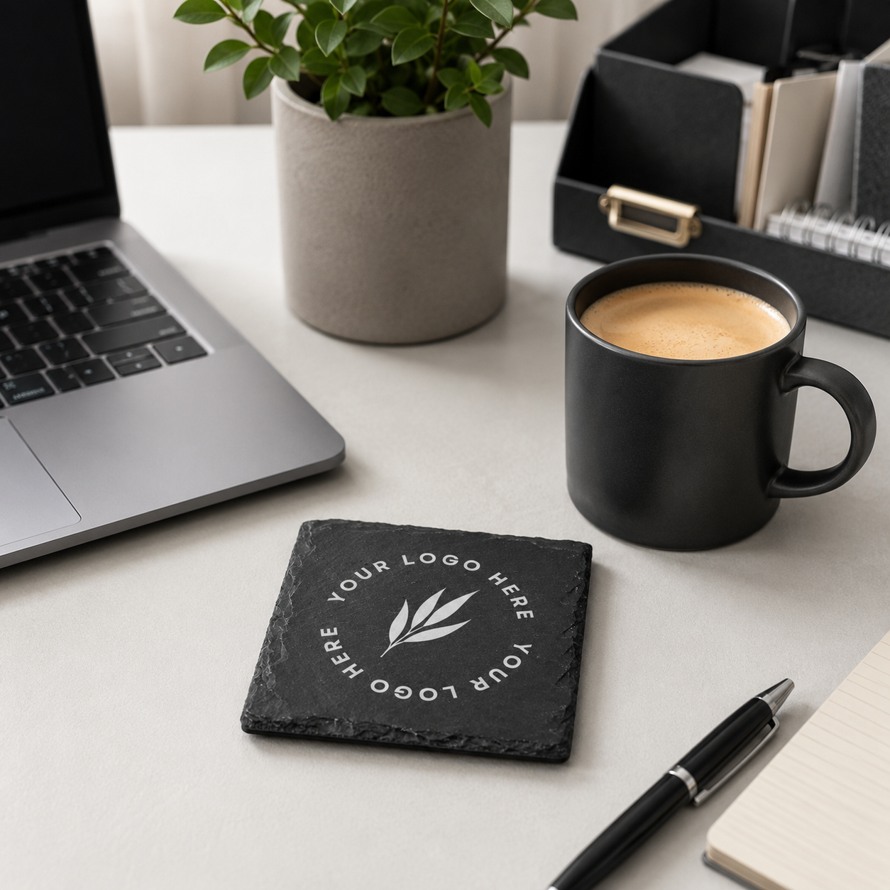 A square black slate coaster with a custom personalised logo sits on a light grey desk next to a black coffee mug, laptop, plant, and pen.