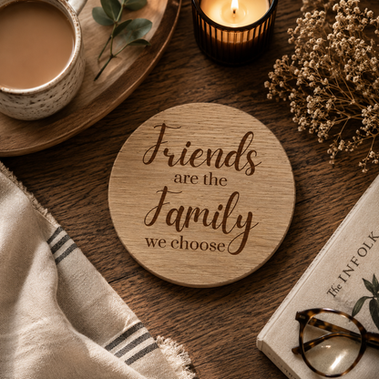 Wooden coaster with 'Friends are the family we choose' text on a wooden table with a cup of coffee, candle, and book.