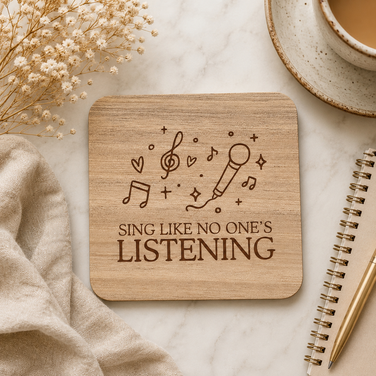 Wooden coaster with 'Sing Like No One's Listening' text on a marble surface with a cup of coffee and notebook.