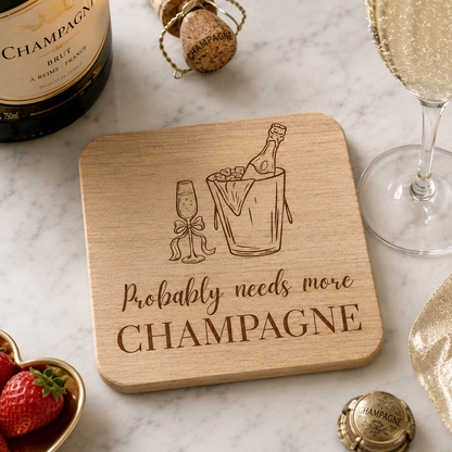 Square wooden coaster featuring the funny quote "Probably needs more champagne" in crisp lettering with a champagne bottle and glass illustration, on a marble bar with champagne and strawberries.