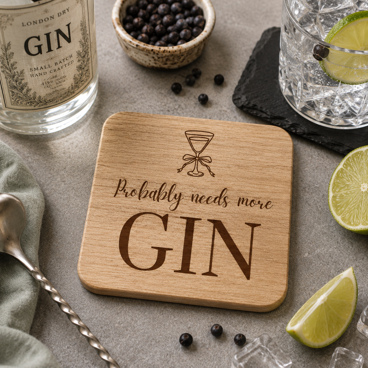  wooden coaster featuring the funny quote "Probably needs more gin" in crisp lettering with a gin glass illustration, on a bar with gin glasses and cocktail accessories.