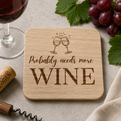 Square wooden coaster with 'Probably needs more wine' text and wine glass illustration, on a marble bar with wine glass, corkscrew and grapes.