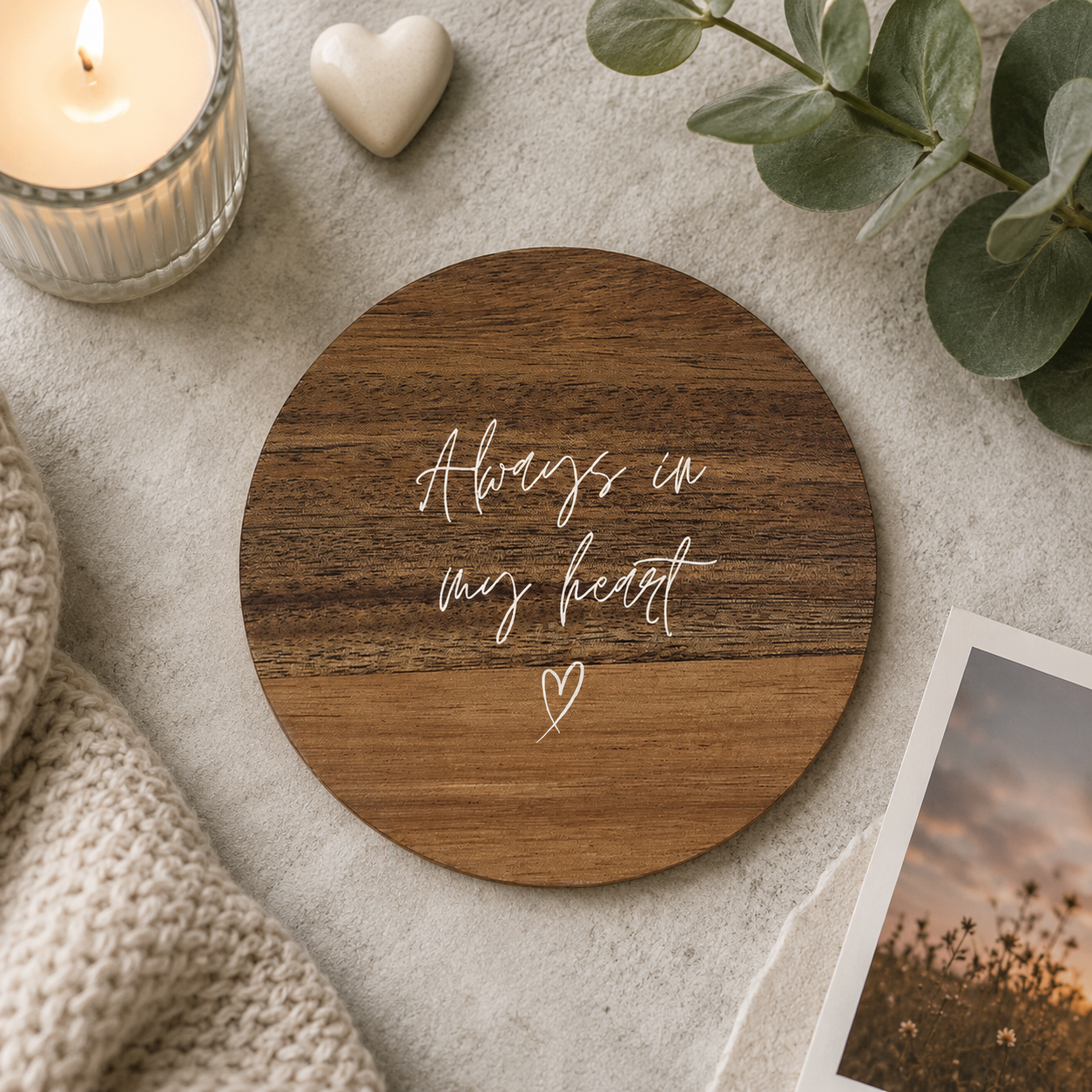 Round wooden coaster with the memorial quote 'Always in my heart' in white lettering, on a stone surface with candles and photos.