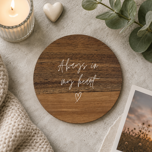 Round wooden coaster with the memorial quote 'Always in my heart' in white lettering, on a stone surface with candles and photos.