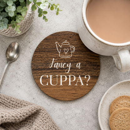 Round wooden coaster with 'fancy a cuppa?' white text and teapot illustration, on a stone surface with cup of tea and biscuits.