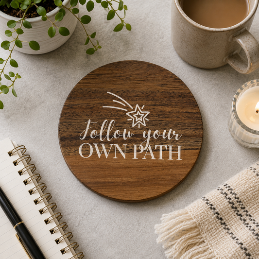 Round wooden coaster with 'follow your own path' white text and shooting star illustration, on a stone surface with tea cup and notebook.