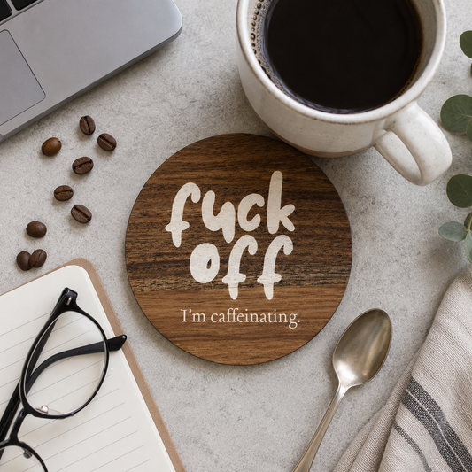 Square wooden coaster featuring the humorous quote "Fuck off I'm caffeinating" in crisp white lettering, on a stone surface with laptop, coffee and glasses.