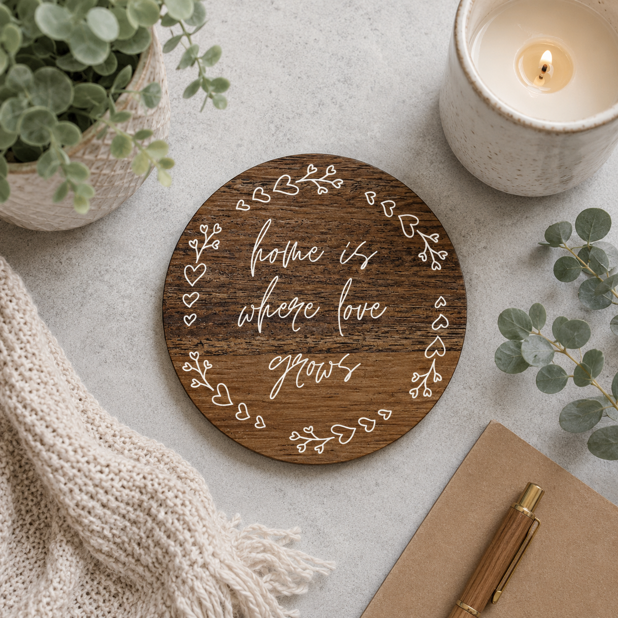 Wooden coaster with 'home is where love grows' text on a table with a candle, plant, and pen.