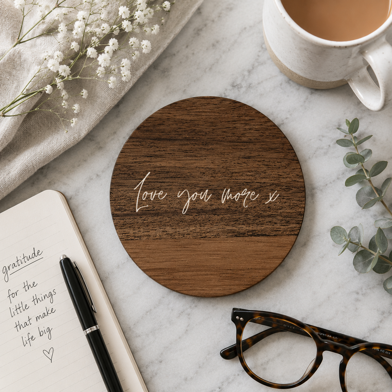 Round wooden coaster with 'Love you more' text on a marble surface with a cup, notebook, and glasses.