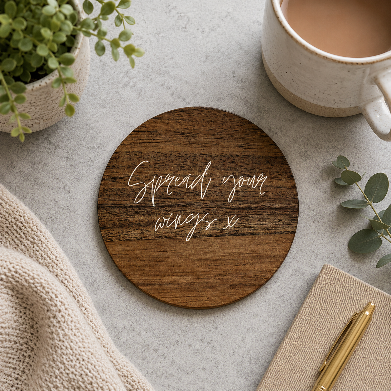 Round wooden coaster with 'Spread your wings' text on a table with a mug, plant, and notebook.