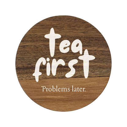 Round wooden coaster with 'Tea first, Problems later.' text on a white background
