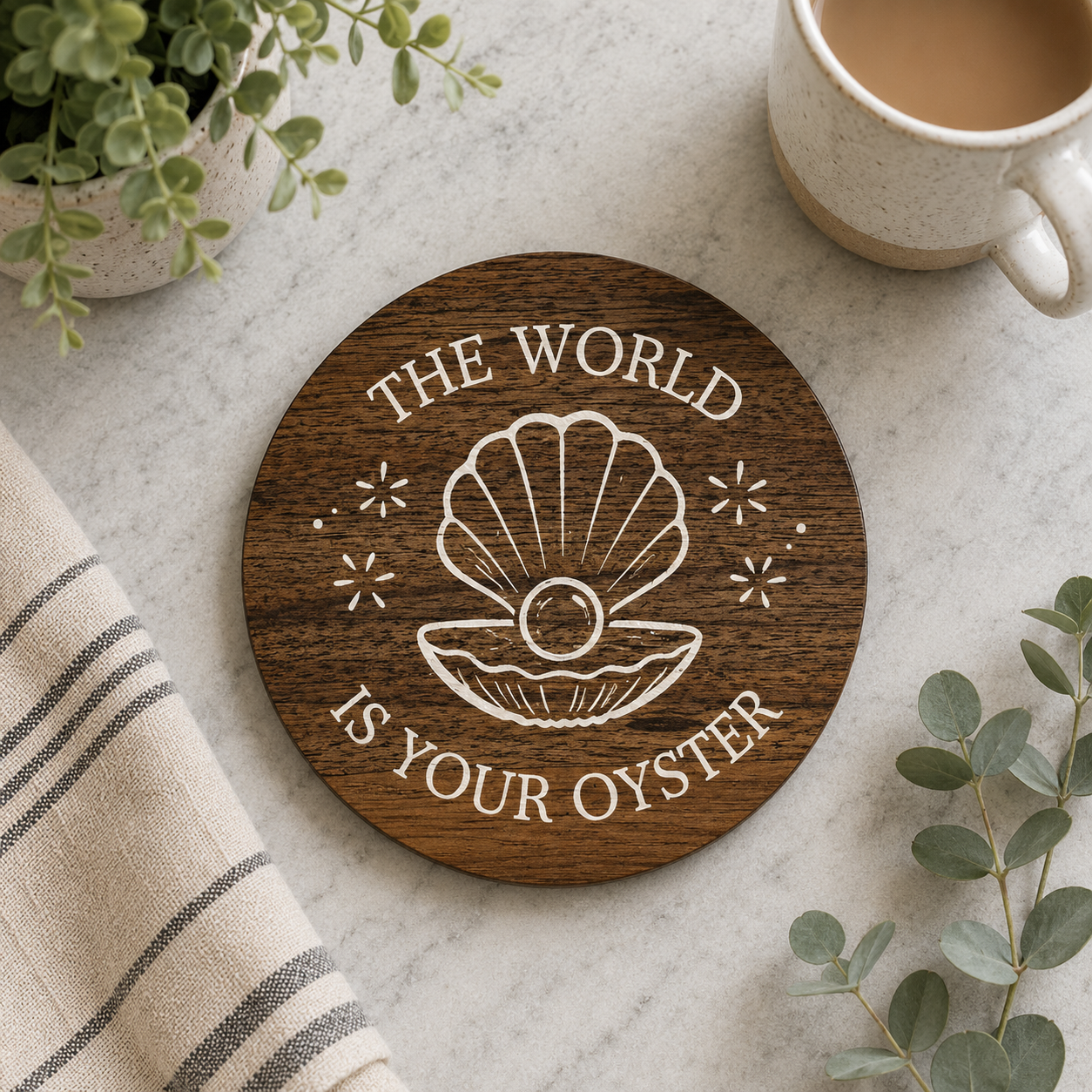 Round wooden coaster with 'The World is Your Oyster' quote on a marble surface with a cup of coffee and plants.