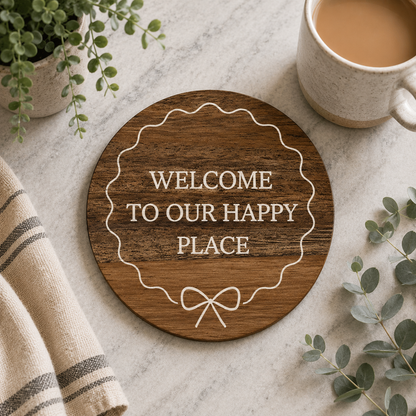 Round wooden coaster with 'Welcome to our happy place' text on a marble surface with a cup of coffee and plants.
