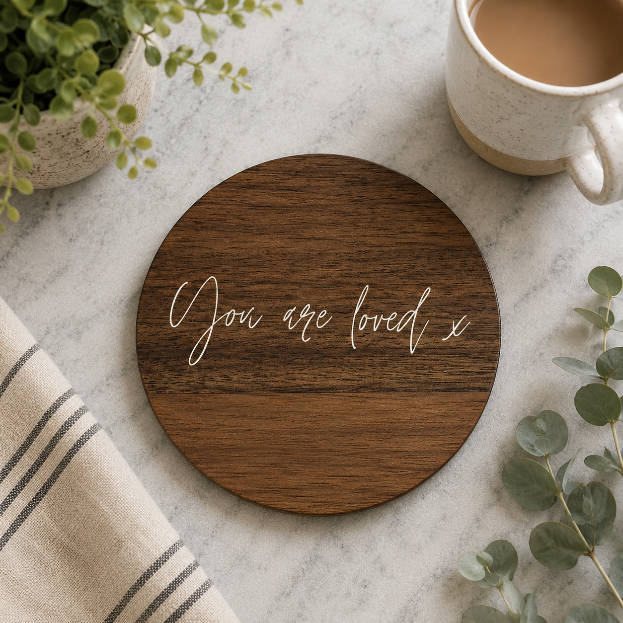 Round wooden coaster with 'You are loved x' text on a table with a cup of coffee and plant.