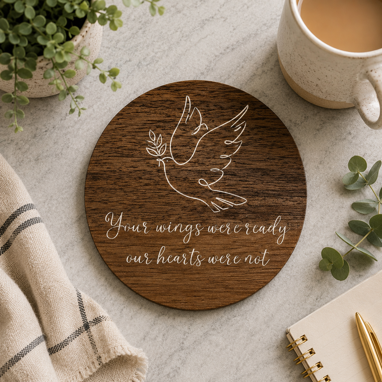Round wooden memorial coaster with a dove design and 'your wings were ready our hearts were not' text on a table with a mug, plant, and notebook.