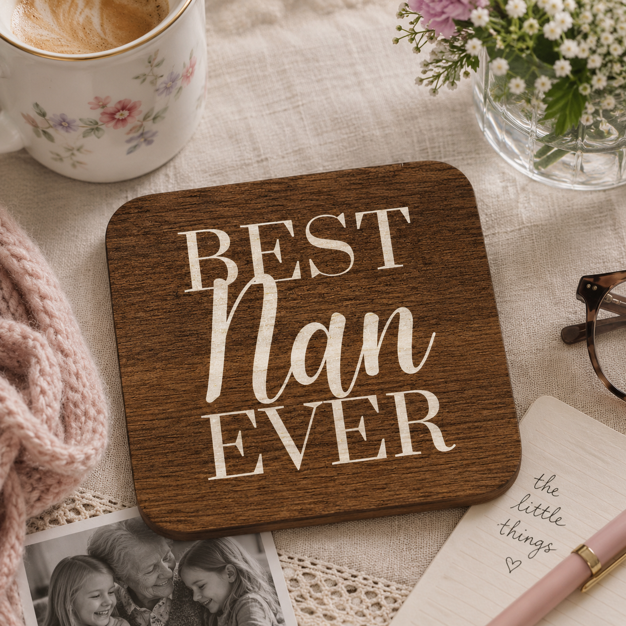 Square wooden coaster with 'Best Nan Ever' text on a table with a cup, flowers, and photos.