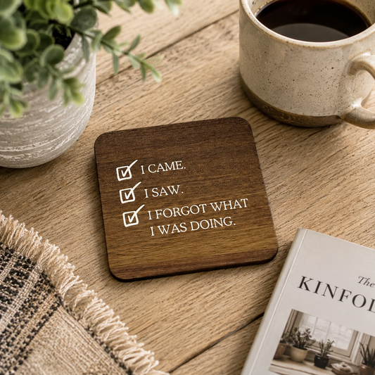 Square wooden coaster with humorous quote 'I came. I saw. I forgot what I was doing.' on a wooden table with a mug and plant.