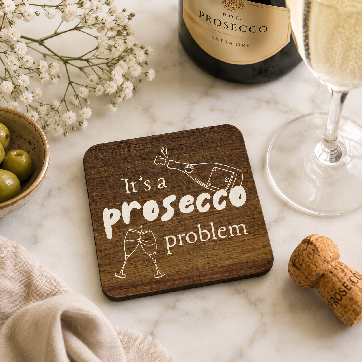 Square wooden coaster with 'It's a prosecco problem' quote on a marble bar with prosecco bottle and glasses.