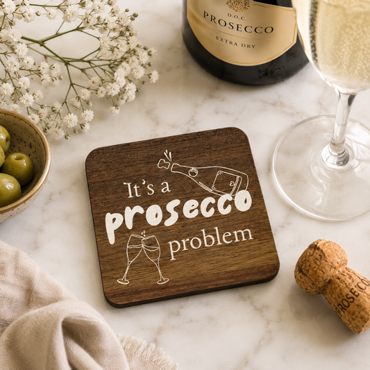 Square wooden coaster with 'It's a prosecco problem' quote on a marble bar with prosecco bottle and glasses.