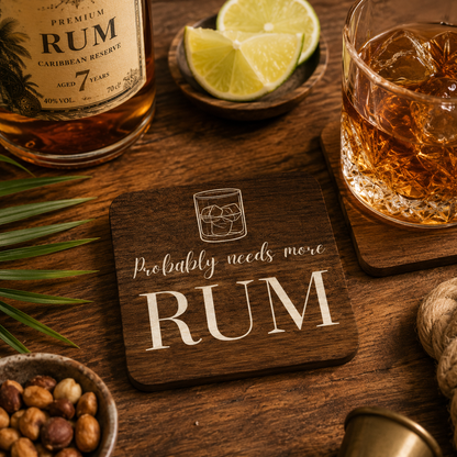 Square wooden coaster with 'Probably needs more rum' text next to a bottle of rum, glass of rum, and lime wedges on a wooden surface.