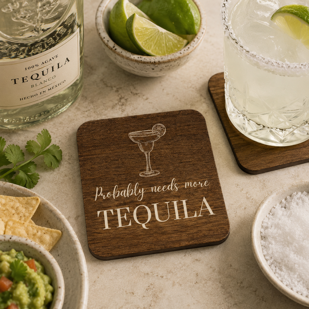 Square wooden coaster with 'probably needs more tequila' quote and margarita glass illustration, on a marble bar with tequila bottle, glass and lime wedges.
