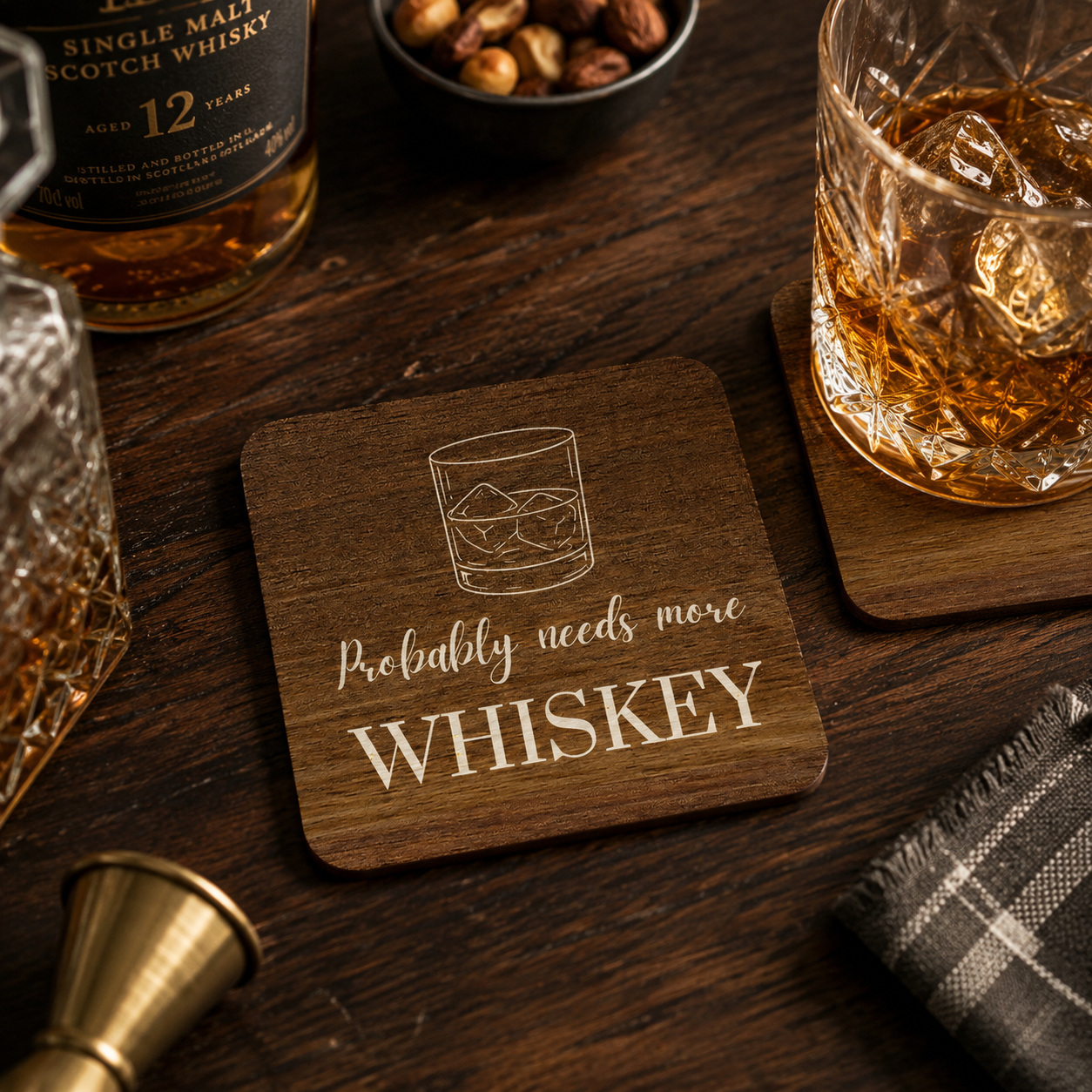 Square wooden coaster with 'probably needs more whiskey' quote and whisky glass illustration, on a wooden bar with whiskey bottle and glass with ice.