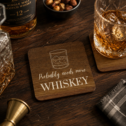 Square wooden coaster with 'probably needs more whiskey' quote and whisky glass illustration, on a wooden bar with whiskey bottle and glass with ice.