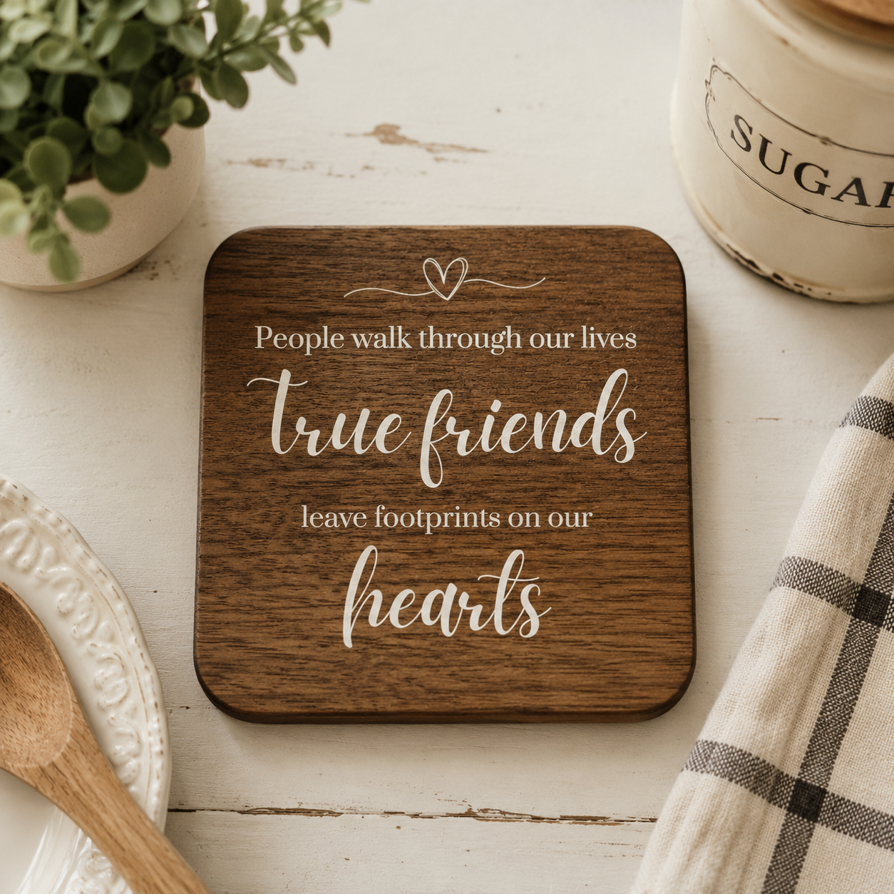 Square wooden coaster with quote 'People walk through our lives, true friends leave footprints on our hearts' in white text, on a kitchen worktop with baking accessories.