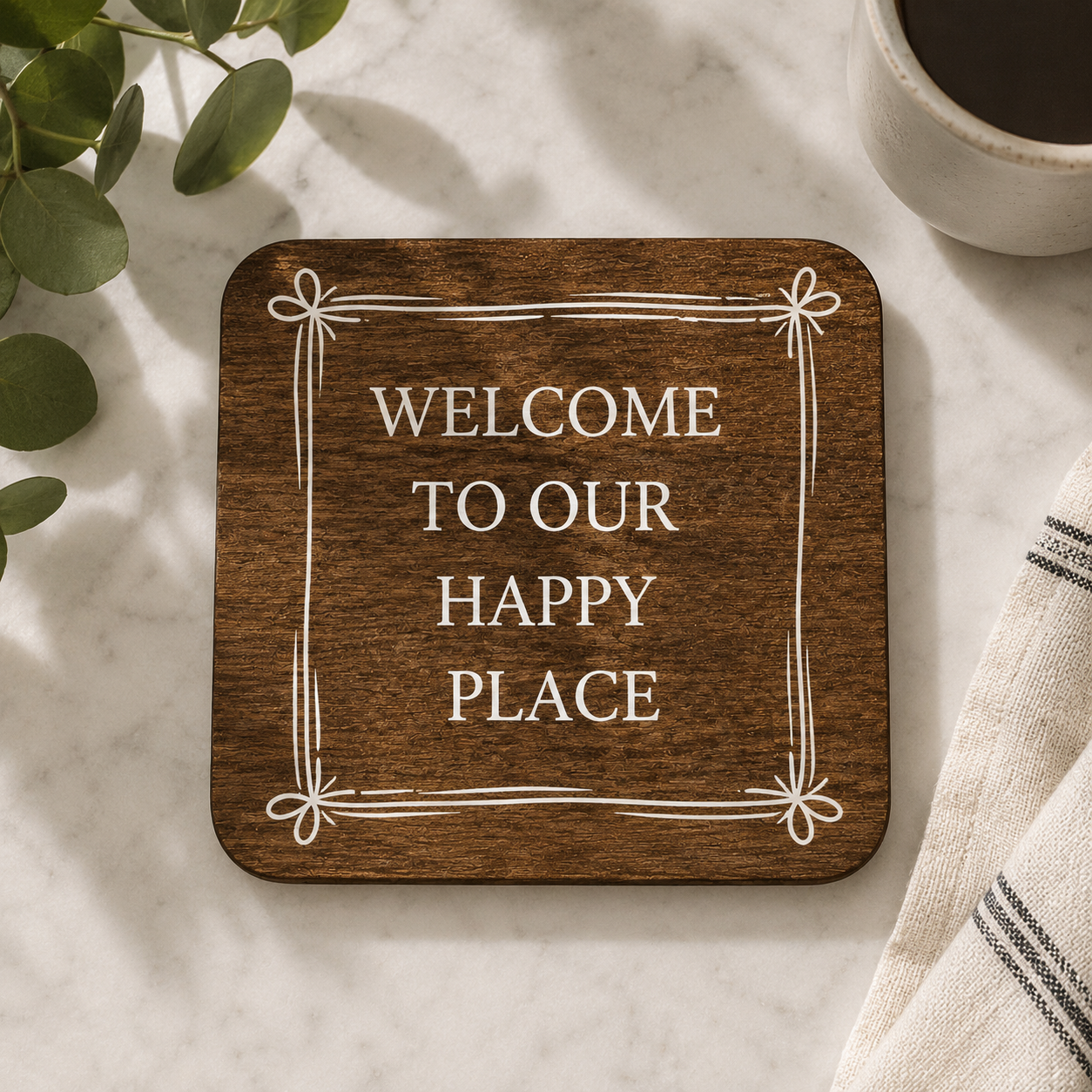 Square wooden coaster with 'welcome to our happy place' quote on a marble work top with coffee cup and plants.