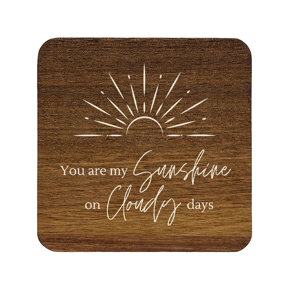 Square wooden coasters with 'you are my sunshine on cloudy days' quote in white text with stylised sunrise illustration.