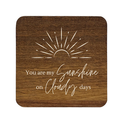 Square wooden coasters with 'you are my sunshine on cloudy days' quote in white text with stylised sunrise illustration.