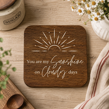 Square wooden coasters with 'you are my sunshine on cloudy days' quote in white text with stylised sunrise illustration, on a wooden work top with flowers and kitchen accessories.