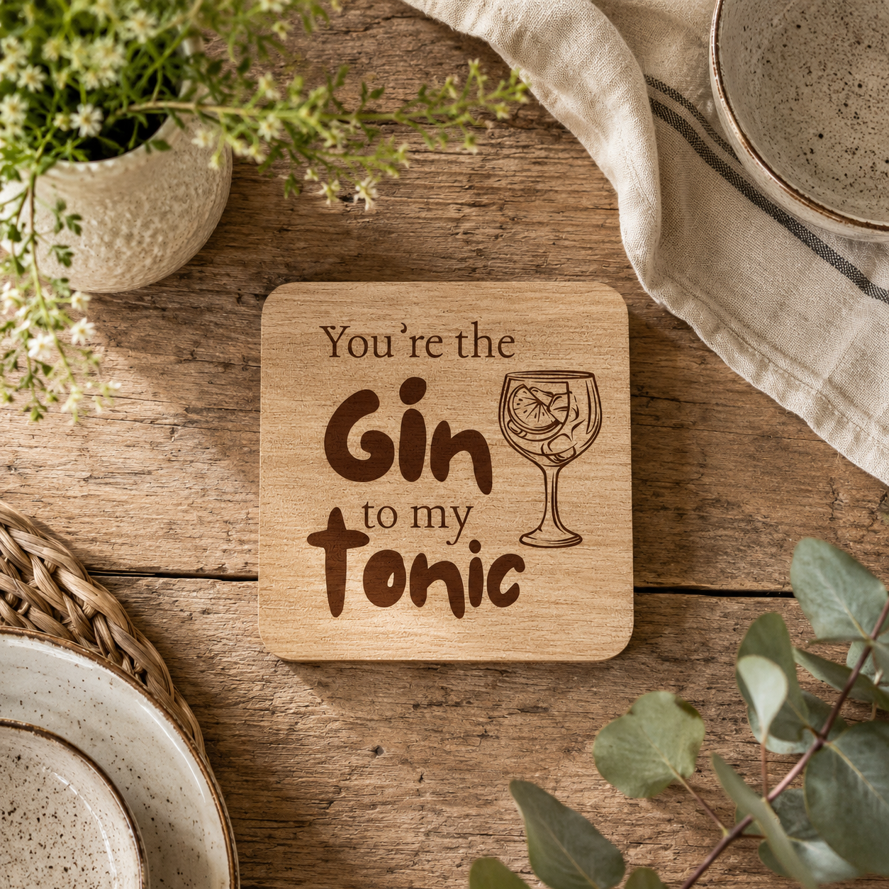 A square wooden coaster with the text 'You're the Gin to my Tonic' and a cocktail glass illustration, styled on a rustic wooden table with plants, plates, and a cloth.