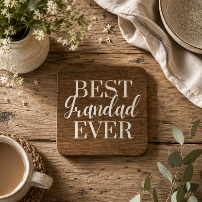 A square wooden coaster with rounded corners reading 'BEST Grandad EVER' in white lettering, styled on a rustic wooden tabletop with flowers, a cup, a cloth napkin, and a bowl nearby.