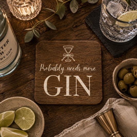 A square wooden coaster with rounded corners sits on a dark wooden table, printed with a cocktail glass icon and the phrase 'Probably needs more GIN' in white lettering. The coaster is styled in a bar scene with a bottle of gin, a glass of gin with a lime, olives, lime slices, a jigger, and a cloth napkin.
