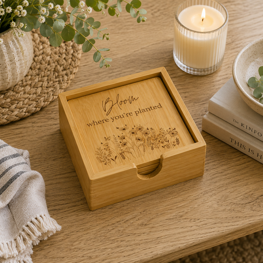 A set of 4 bamboo wood coasters decorated with inspirational quotes and wildflower illustrations. The coasters are stored in a matching bamboo box on a wooden coffee table next to a candle and books.