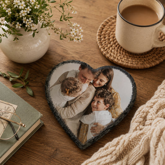 Heart-shaped personalised slate coaster with a photo of a family on a wooden table with a cup of coffee, book, and flowers.