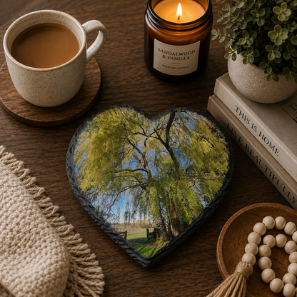 Heart-shaped personalised photo slate coaster with tree photo, cup of coffee, candle, and books on a wooden surface.