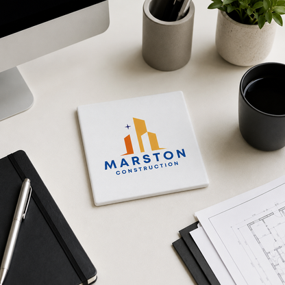 A square white coaster featuring ta custom personalised business logo, styled on a light beige desk with a computer monitor, notebook, pen, coffee cup, and plant.