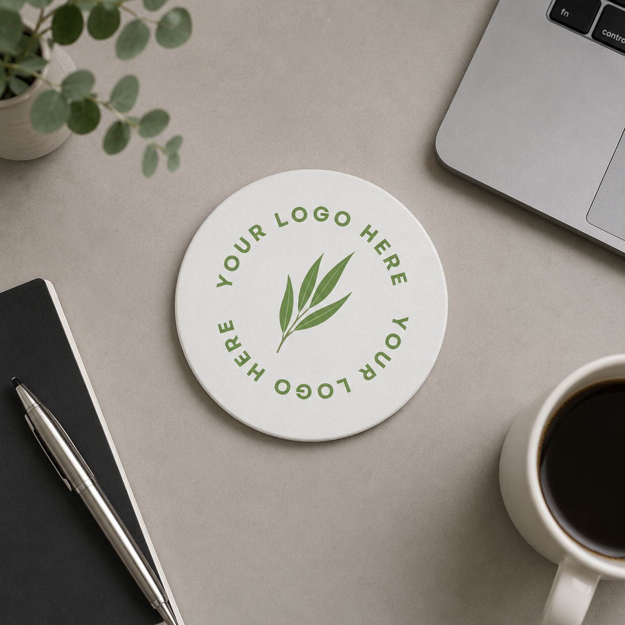 A round white coaster with a custom business logo logo, shown in a styled flat-lay on a beige desk with a laptop, notebook, pen, plant, and coffee cup.