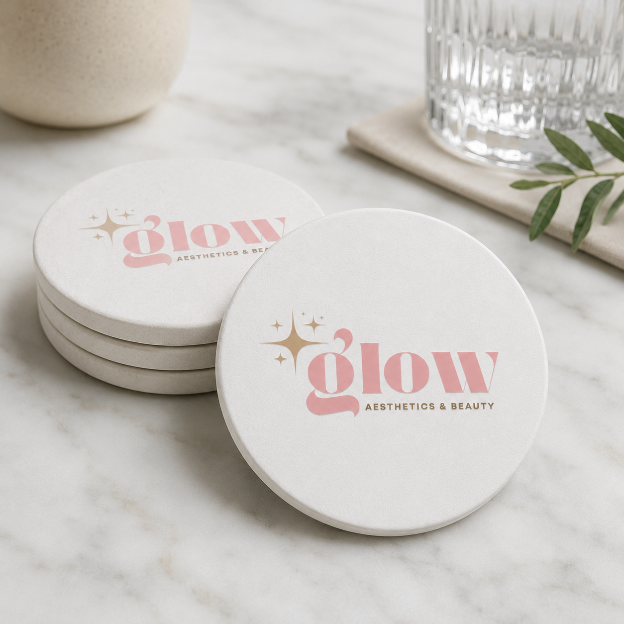 Four round white coasters stacked on a marble surface, with the front coaster showing a personalised business logo. A glass, a ceramic bowl, and green leaves are softly blurred in the background.