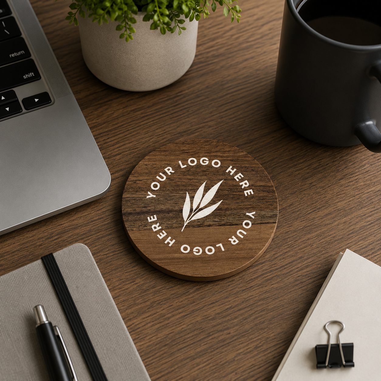 A round wooden desk coaster with a white printed leaf logo and the text 'YOUR LOGO HERE' in a circular arrangement, shown on a wooden desk surrounded by a laptop, notebook, pen, plant, and a black mug.