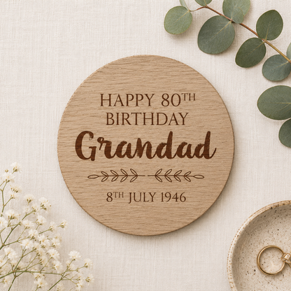 Personalised wooden round coaster with 'Happy 80th Birthday Grandad' text on a light background with flowers and a ring.
