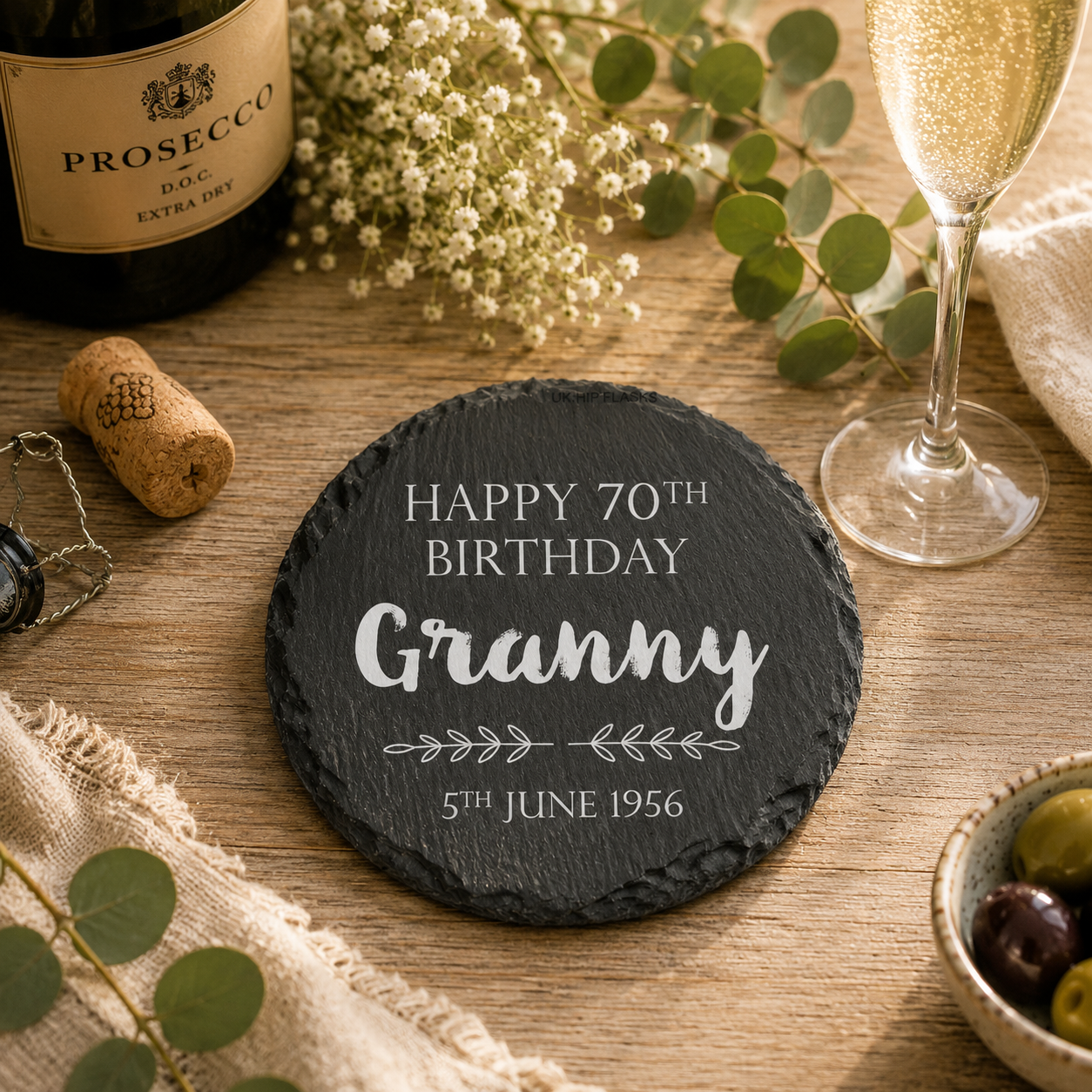 Personalised round slate coaster with 'Happy 70th Birthday Granny' text on a wooden table with Prosecco and flowers.