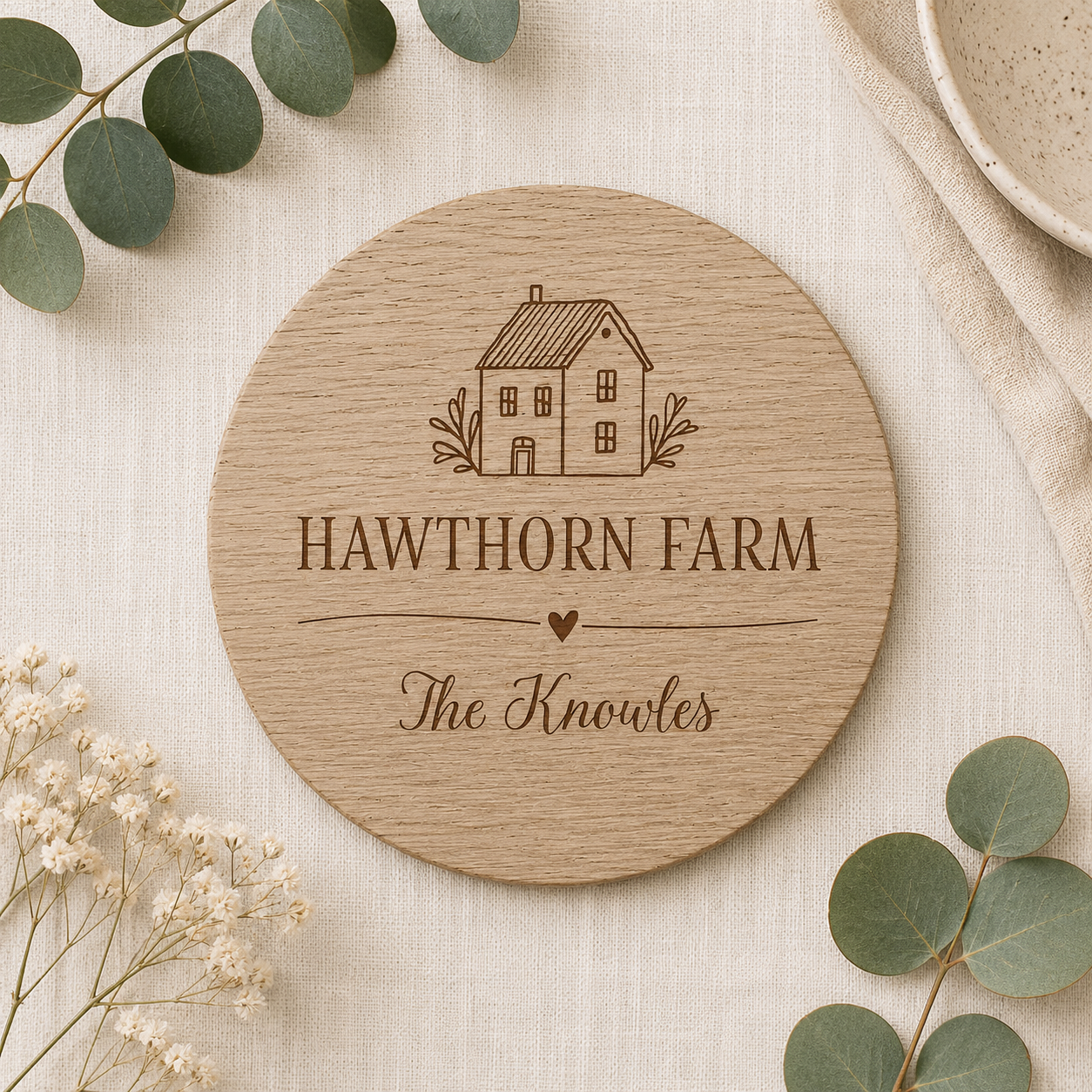 Round wooden coaster with personalised house and family name on a light background with greenery.