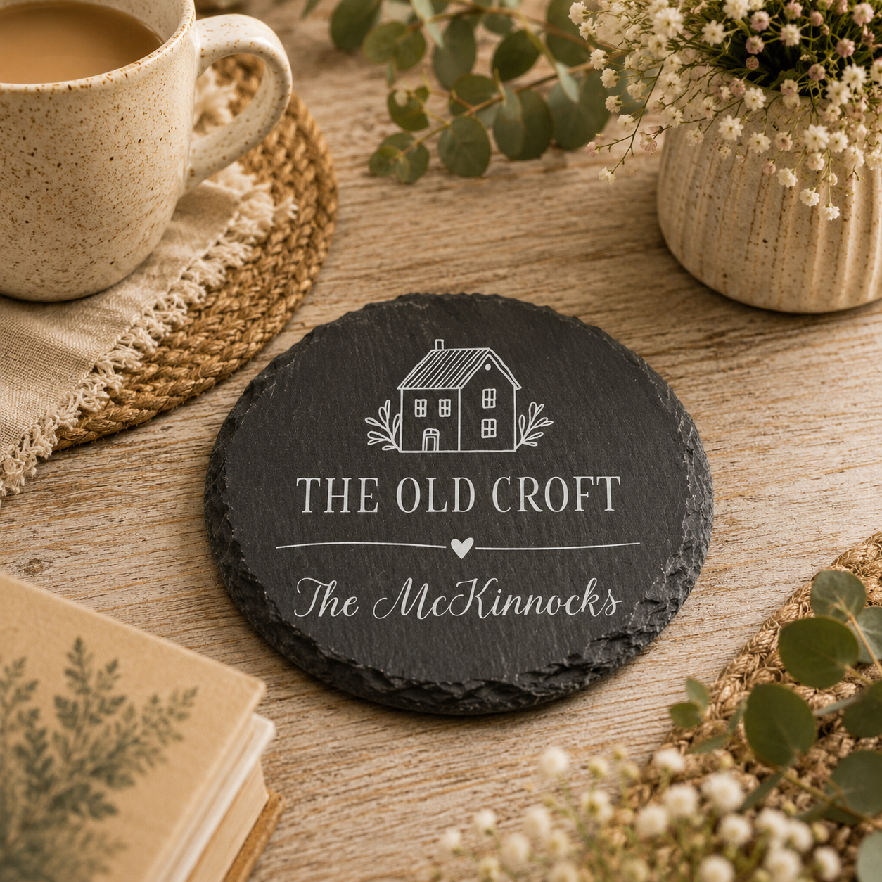 Round personalised house name or number slate coaster, personalised with a house name and family name. The coaster is on a wooden surface with a cup of tea, plants and books.