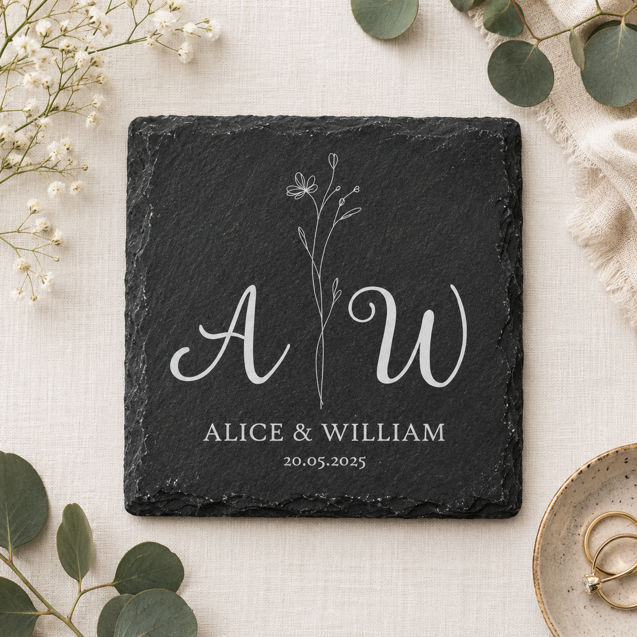 Square slate coaster personalised with a couple's initials, names and date, on a fabric surface with plants and wedding rings.