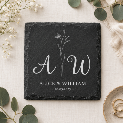 Square slate coaster personalised with a couple's initials, names and date, on a fabric surface with plants and wedding rings.