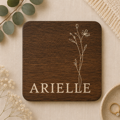 Square wooden coaster with personalised name and floral design on a beige surface with decorative elements.